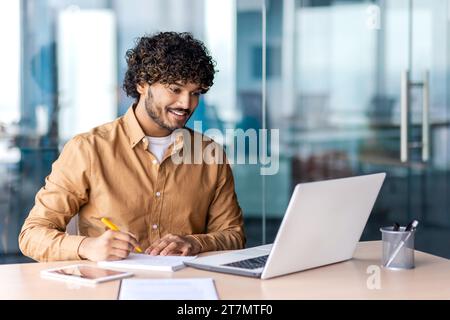 Man studying online remotely, businessman improving qualifications at the workplace inside office, writing down data in a notebook, watching a video course, satisfied with high results, smiling. Stock Photo