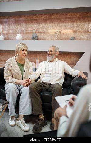 middle aged married couple sitting on leather couch near psychologist taking notes during consult Stock Photo