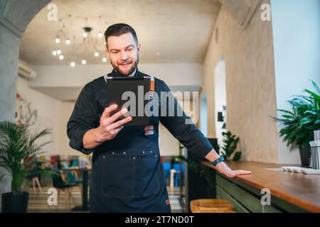 Smiling small business owner dressed in a black chef uniform with an apron using digital tablet in his cozy restaurant hall. Successful people, hard w Stock Photo