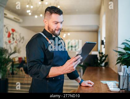 Smiling small business owner dressed in a black chef uniform with an apron using digital tablet in his cozy restaurant hall. Successful people, hard w Stock Photo