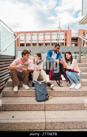 Vertical. Group of multiracial high school students talking on a staircase at university campus, using a tablet app and smartphone to do homework project after class. Friendly teenage people studying. High quality photo Stock Photo