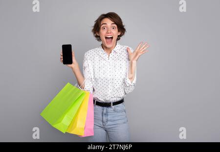 Excited cute woman with shopping bags shows her smart phone and shouting. Black Friday, consumer, buyer, sale, store, payment, online buying, discount Stock Photo