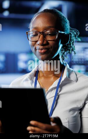 Satisfied admin in data center uses tablet to prevent system electronics overload. Efficient expert between server mainframes ensuring enough network bandwidth for smooth operations Stock Photo