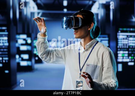 Admin in data center uses virtual reality to prevent system overload during peak traffic periods. Man with VR headset in server farm ensuring enough network bandwidth for smooth operations Stock Photo