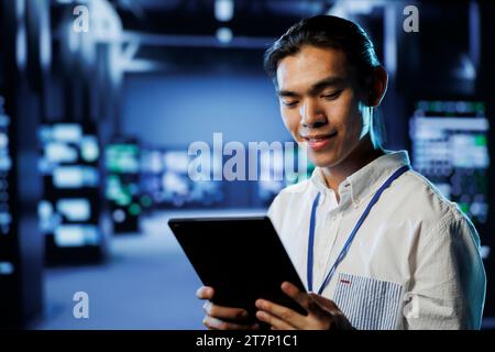 Cheerful computer operator in data center uses tablet to prevent system overload during peak traffic periods. BIPOC man in server facility ensuring enough network bandwidth for smooth operations Stock Photo