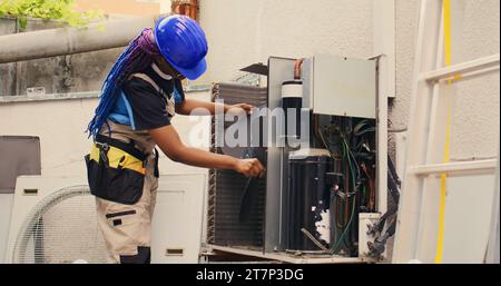 Certified technician commissioned by customer for annual air conditioner routine maintenance, using soft dusting brush to clean dusty condenser coils and filters, outdoor Stock Photo