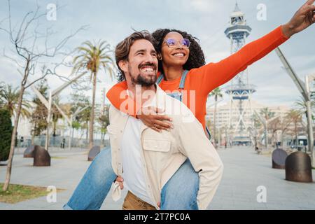 Young smiling adult man carrying on shoulders his joyful girlfriend while laughing and having fun together outside. Portrait of multiracial couple giving a piggyback ride, relaxing and enjoying. High quality photo Stock Photo
