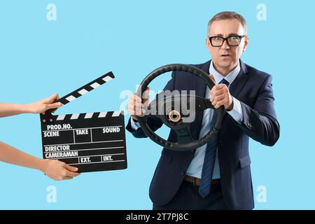 Mature actor holding steering wheel and hands with clapperboard on blue background Stock Photo