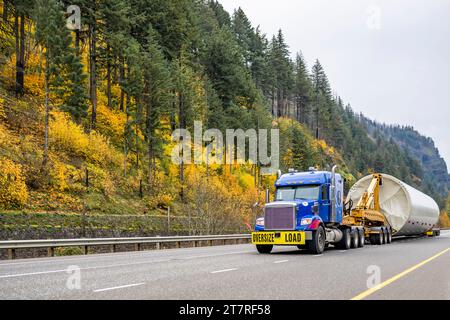 Industrial carrier big rig blue semi truck tractor with oversize load sign transporting part of oversized wind turbine pillar on semi trailer with tro Stock Photo