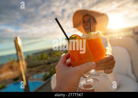 Close-up of a tropical cocktail with a straw held by a woman in a sun hat at tropical beach hotel on sunset Stock Photo