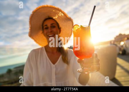 Smiling woman in straw hat holding a cocktail against a sunset backdrop Stock Photo