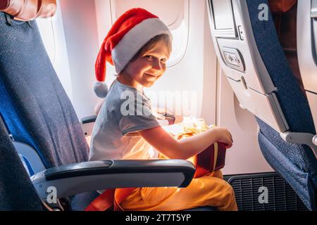 Child in Santa Claus hat flies at cabin of airplane with holiday gift. Stock Photo