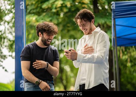 Two, male students watching something funny on the phone and burst out laughing during break at school while standing at the university quad. Stock Photo