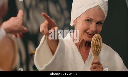Funny mature middle-aged 50s woman in bathrobe sing at hairbrush in bathroom mirror reflection happy old female lady singing song like singer having Stock Photo