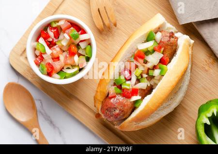 A choripan with Argentinian criolla sauce on a wooden board, a green pepper, wooden fork and spoon, and a brown napkin on white marble background. Stock Photo