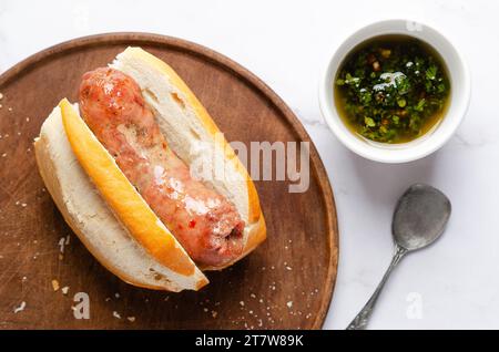 A choripán on a wooden board, a bowl with chimichurri and a spoon on white marble background. Stock Photo