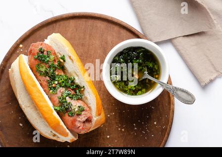 A choripan with chimichurri sauce on a wooden board and a brown napkin on white marble background. Stock Photo