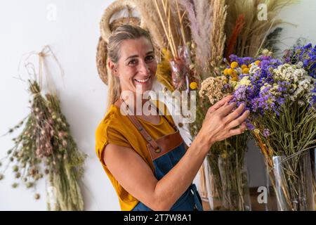 joyful florist woman in a mustard-colored shirt and denim apron holds a bouquet of dried flowers, surrounded by an array of florals against a white ba Stock Photo