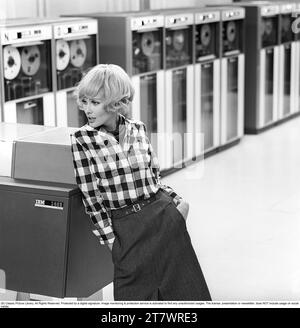 In the 1960s. Fashion model wearing the typical clothes of 1965. The photograph is taken in a 1960s mainframe computer room with magnetic tape units in use where the tape containing information is being read or entered.  She is wearing a chequered shirt. Picture taken 1965. ref DV16 Stock Photo