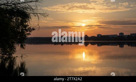 Scenic warm sunrise on cloudy sky reflecting in calm lake water. Peaceful morning in city park by the river Stock Photo