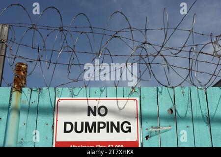 No dumping sign on fence with barbed wire and razor wire against blue sky. Stock Photo