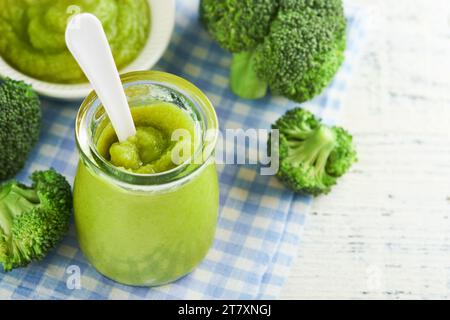 Green broccoli baby food in white bowl and jar on table. Green baby food. Child first feeding concept. Baby Natural Food. Production and menu of baby Stock Photo