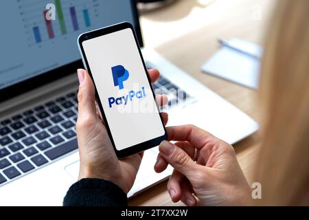 Paypal app on the screen of smartphone. Woman in front of a laptop with mobile phone in her hands. Logotype of electronic payment system. Rosario, Arg Stock Photo