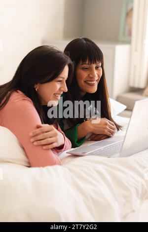 Happy biracial mother and teenage daughter lying on bed and using laptop at home, copy space Stock Photo