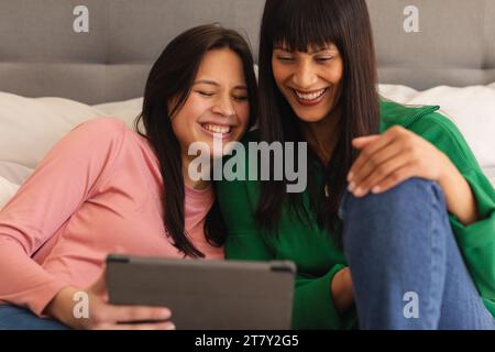 Happy biracial mother and teenage daughter on bed and using tablet at home, copy space Stock Photo