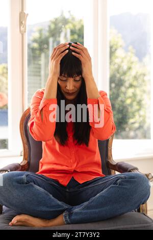 Upset biracial woman having video call sitting cross legged in sunny room holding head Stock Photo
