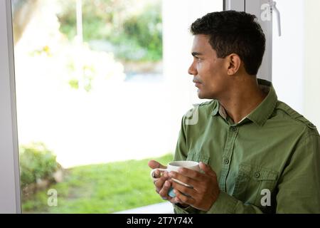 Thoughtful biracial man holding mug looking through window at home Stock Photo