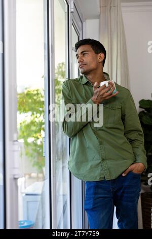 Thoughtful biracial man holding mug looking through window at home Stock Photo