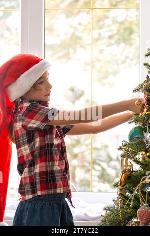 Happy girl in a Santa hat decorates the Christmas tree with colorful figurines. The child is having fun barefoot, in a dwarf's hood near the window, w Stock Photo