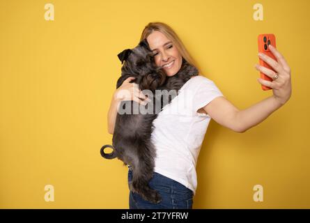 Friendship concept. Happy european woman taking selfie with her cute schnauzer dog, looking and smiling at camera over yellow studio background wall Stock Photo