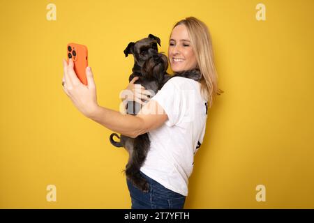 Friendship concept. Happy european woman taking selfie with her cute schnauzer dog, looking and smiling at camera over yellow studio background wall Stock Photo