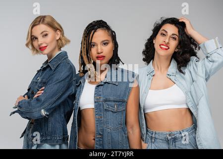 fashionable multiracial girlfriends in blue denim clothes looking at camera on grey, diverse beauty Stock Photo