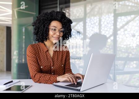 Young female employee programmer working at workplace inside office with laptop, business woman typing on keyboard happily smiling, satisfied with work and achievement results. Stock Photo