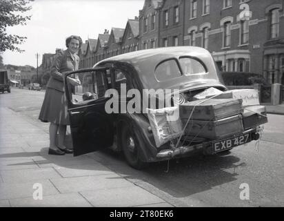 1950s, historical, outside in a suburban street, a lady beside her car of the era, with luggage, travel trunks and suitcases tied down by string on the open flap of the car's boot, England, UK. The boot's on cars made in this era had limited storage space. Also notable on cars made in the 1930s and 40s, are the rear-hinged doors, as seen here. Stock Photo