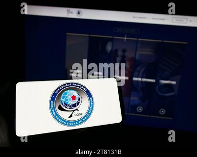 Person holding mobile phone with logo of Direction Generale de la Securite Exterieure (DGSE) in front of web page. Focus on phone display. Stock Photo