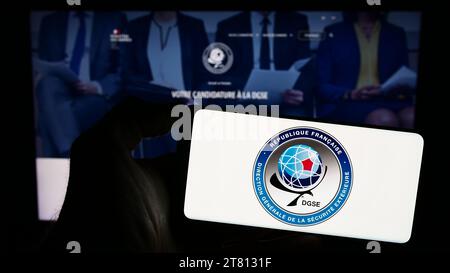 Person holding cellphone with logo of Direction Generale de la Securite Exterieure (DGSE) in front of webpage. Focus on phone display. Stock Photo
