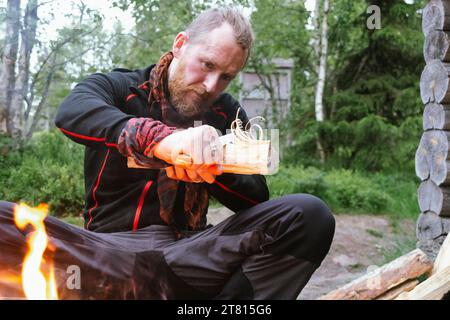 A man carves feather sticks in a piece of wood to start a fire in the forest on a hiking trail in northern Finland Stock Photo