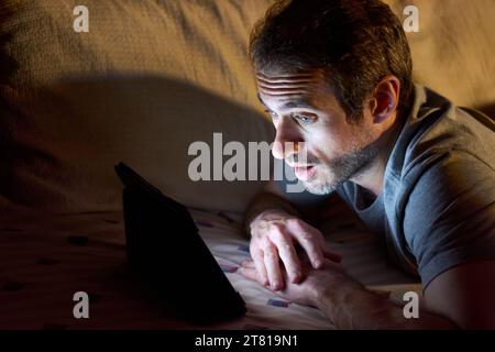 Young 30 year old man lying in bed at night while watching something on a tablet with expression of surprise on his face. Copy space for text. Stock Photo