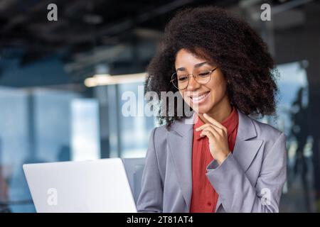 Young beautiful successful business woman close up smiling looking at laptop screen, female worker in business suit with curly hair is reading contentedly, woman is happy results of achievement. Stock Photo