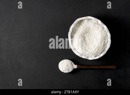 natural coconut flakes in the bowl of shape of coconut with spoon on black table, copy space. Stock Photo