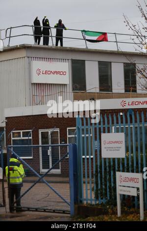 Southampton, Hampshire, UK. 16th Nov, 2023. Activists raise their fists in the air from on top of the roof as a security guard stands-by at the gate. Palestine Action occupy the roof of Italian arms industry giant Leonardo at their in their factory in Southampton and shuts them down. Leonardo supply Israel with fighter jets and weaponry that are currently being used in Gaza. Palestine Action demands that arms companies providing weapons for Israel shut down permanently. They have announced that companies selling weapons to the Israeli Defence Force and their partner companies will be target Stock Photo