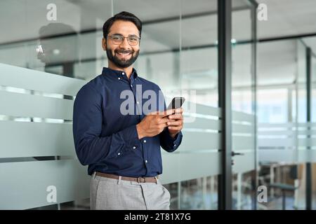 Smiling Indian businessman holding phone standing in office. Portrait. Stock Photo
