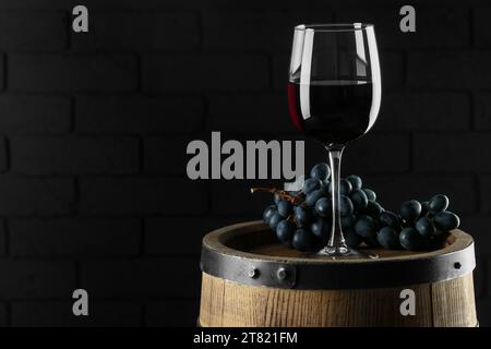 Glass with delicious wine and fresh grapes on wooden barrel against dark wall, space for text Stock Photo