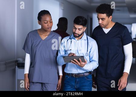 Diverse male and female doctors using tablet walking in hospital corridor Stock Photo