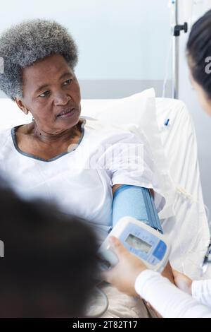 Diverse female doctor testing blood pressure of senior female patient in hospital room Stock Photo