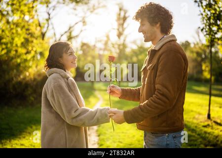 Portrait of happy loving couple in park in sunset. Man is giving rose to his woman. Stock Photo